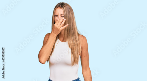 Young blonde woman wearing casual style with sleeveless shirt smelling something stinky and disgusting, intolerable smell, holding breath with fingers on nose. bad smell