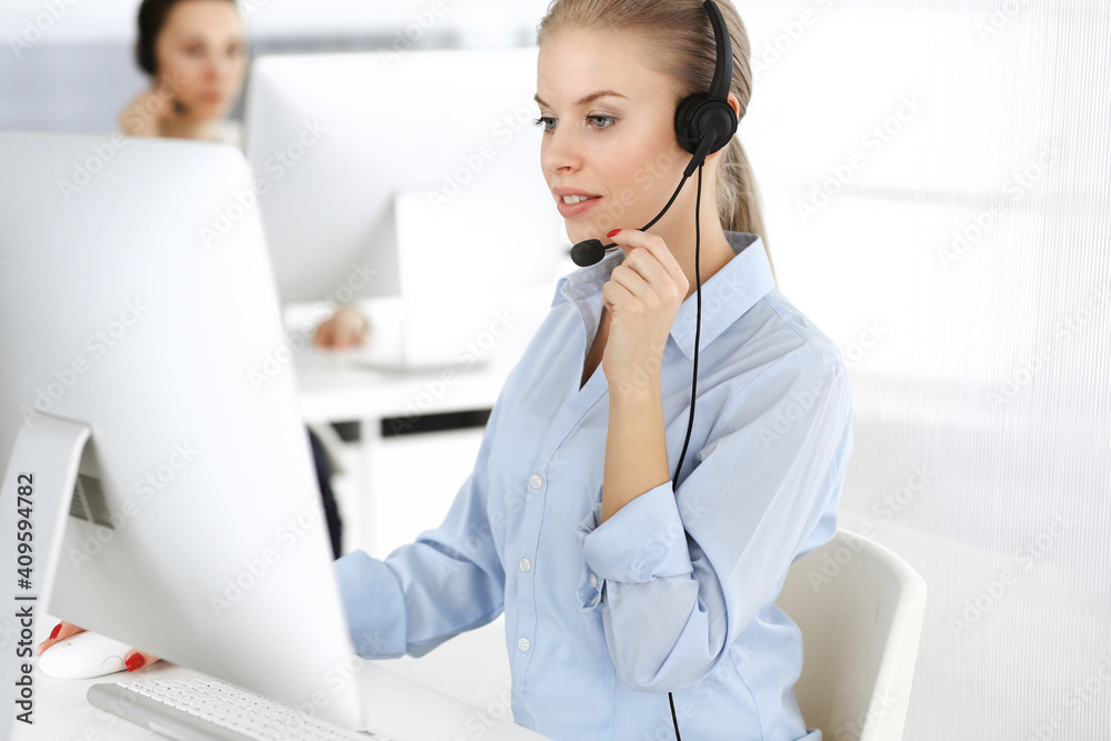 Blond woman call operator is using computer and headset for consulting clients online. Group of diverse people working as customer service occupation. Business concept Blond woman call operator is using computer and headset for consulting clients online. Group of diverse people working as customer service occupation. Business concept