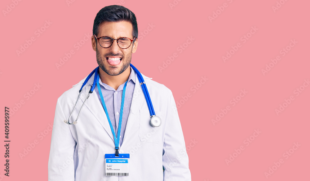 Young hispanic man wearing doctor uniform and stethoscope sticking tongue out happy with funny expression. emotion concept. Young hispanic man wearing doctor uniform and stethoscope sticking tongue out happy with funny expression. emotion concept.