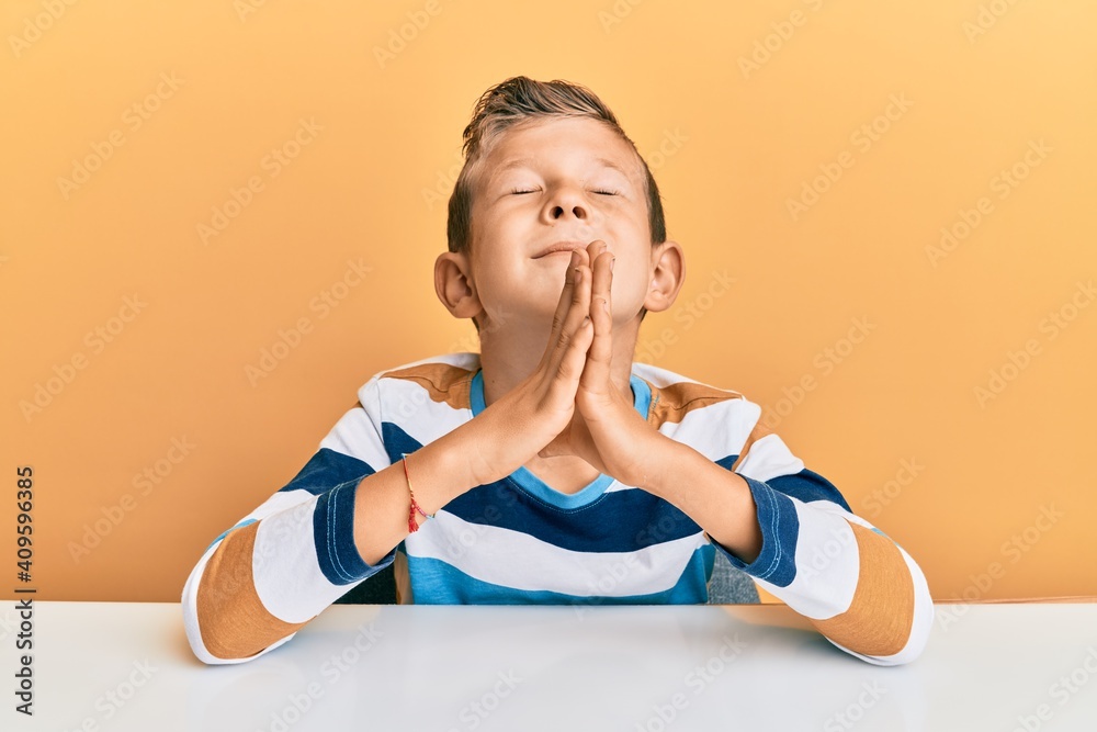 Adorable caucasian kid wearing casual clothes sitting on the table begging and praying with hands together with hope expression on face very emotional and worried. begging. Adorable caucasian kid wearing casual clothes sitting on the table begging and praying with hands together with hope expression on face very emotional and worried. begging.