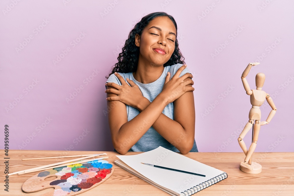 Young african american girl artist sitting at studio table hugging oneself happy and positive, smiling confident. self love and self care Young african american girl artist sitting at studio table hugging oneself happy and positive, smiling confident. self love and self care