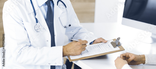 Doctor and patient discussing medical exam results while sitting at the desk in clinic, close-up. Male physician using clipboard for filling up medication history record of young woman