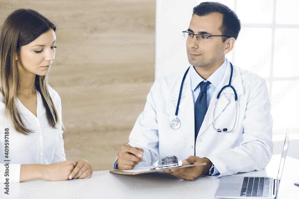Fototapeta premium Doctor and patient discussing medical exam results while sitting at the desk in clinic. Male physician using clipboard for filling up medication history record of young woman. Data in medicine