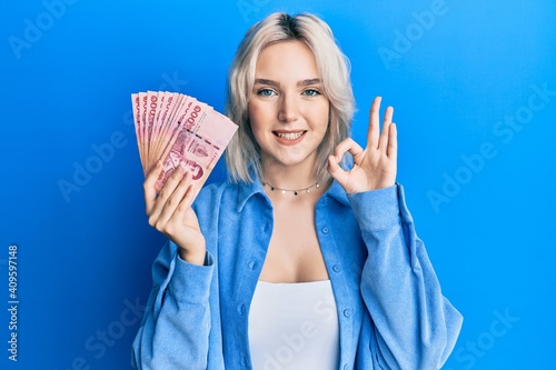 Slika na platnu Young blonde girl holding thai baht banknotes doing ok sign with fingers, smilin