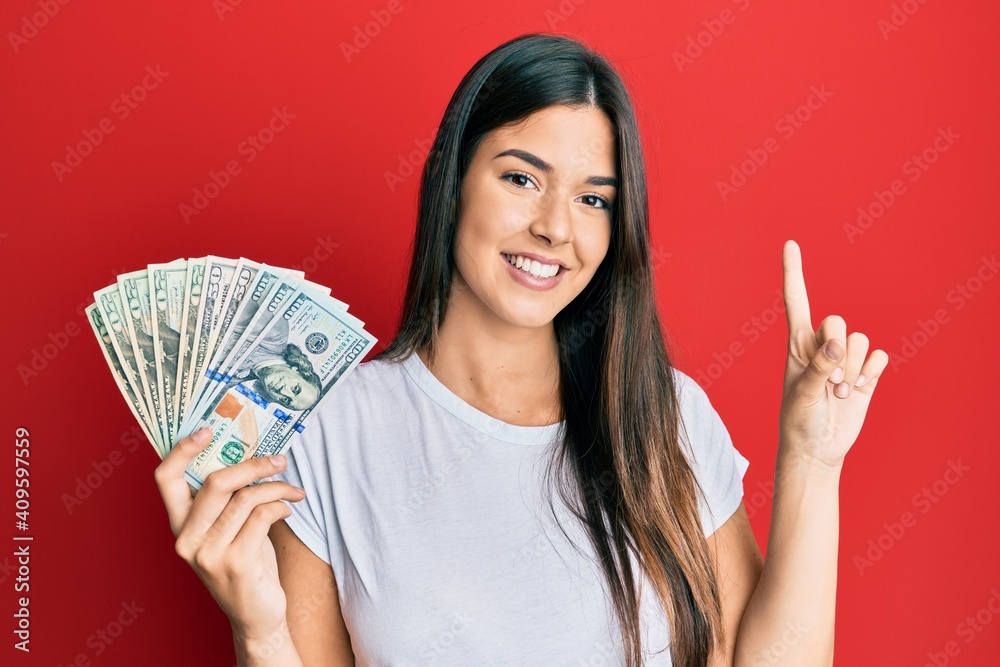 Young brunette woman holding dollars smiling with an idea or question pointing finger with happy face, number one Young brunette woman holding dollars smiling with an idea or question pointing finger with happy face, number one
