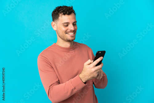 Young caucasian handsome man isolated on blue background sending a message or email with the mobile © luismolinero