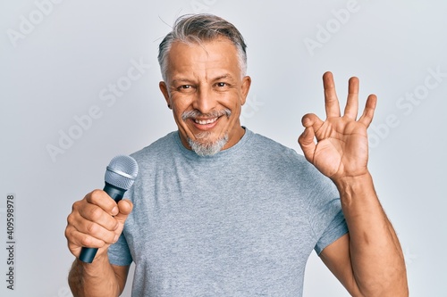 Middle age grey-haired man singing song using microphone doing ok sign with fingers, smiling friendly gesturing excellent symbol