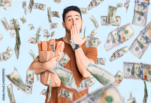 Young hispanic man holding paper bag with bread covering mouth with hand, shocked and afraid for mistake. surprised expression photo