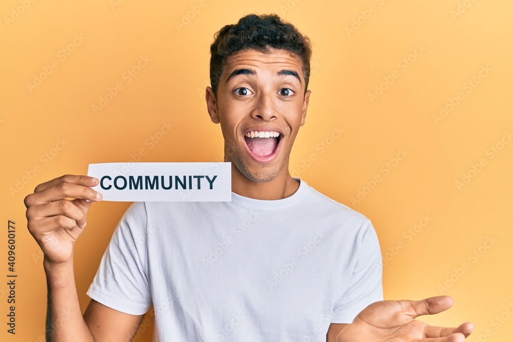Young handsome african american man holding community baner as social cooperation celebrating achievement with happy smile and winner expression with raised hand Young handsome african american man holding community baner as social cooperation celebrating achievement with happy smile and winner expression with raised hand