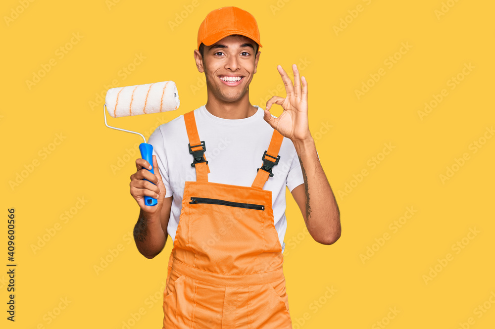 Young handsome african american man wearing cap and painter clothes holding painting roll doing ok sign with fingers, smiling friendly gesturing excellent symbol
