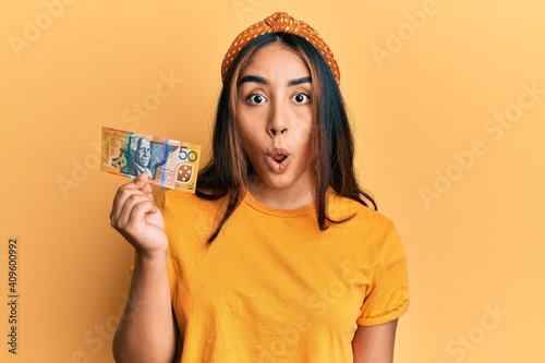 Young latin woman holding 50 australian dollar banknote scared and amazed with open mouth for surprise, disbelief face