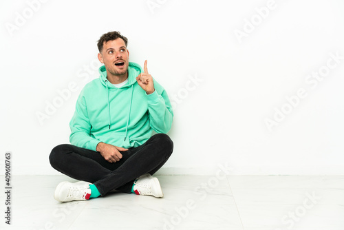 Young handsome caucasian man sitting on the floor thinking an idea pointing the finger up