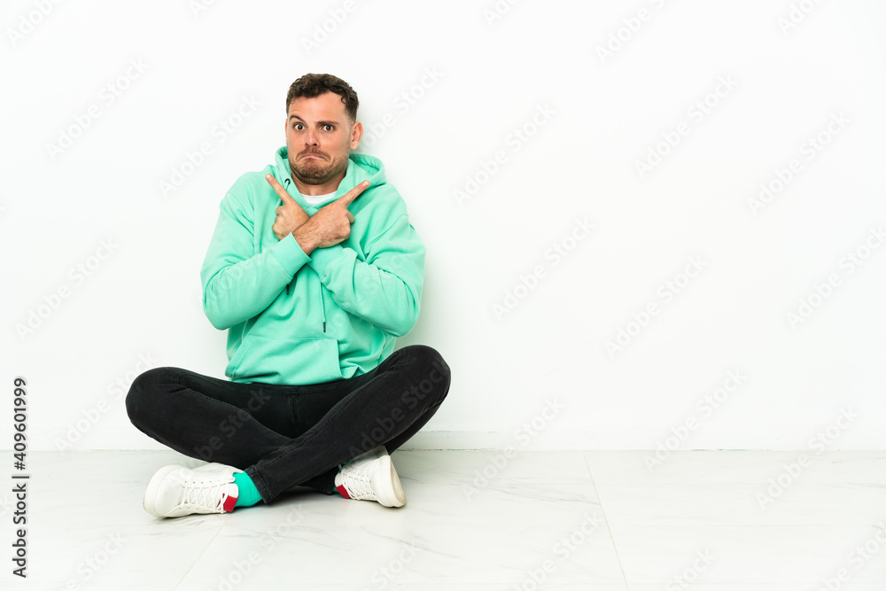 Young handsome caucasian man sitting on the floor pointing to the laterals having doubts Young handsome caucasian man sitting on the floor pointing to the laterals having doubts