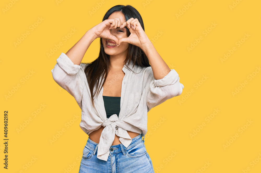 Young beautiful hispanic girl wearing casual clothes doing heart shape with hand and fingers smiling looking through sign Young beautiful hispanic girl wearing casual clothes doing heart shape with hand and fingers smiling looking through sign