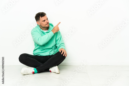 Young handsome caucasian man sitting on the floor pointing back with the index finger