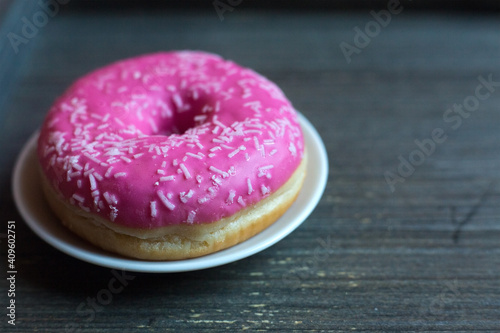 side view to bright pink donut on white round plate and aged wooden grey background. Isolated object close up. Traditional American sweets for breakfast. Copy space. White sugar decoration on top.  photo