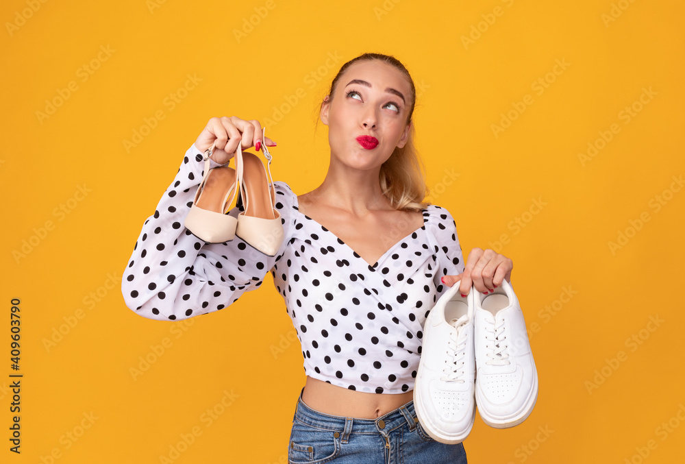 Pensive woman holding pair of high heels shoes and sneakers Pensive woman holding pair of high heels shoes and sneakers
