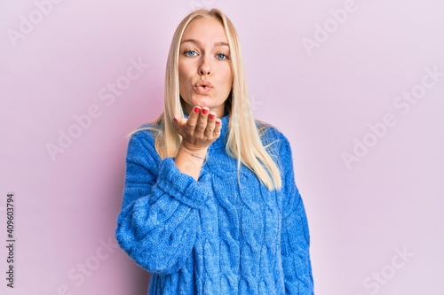 Young blonde girl wearing wool winter sweater looking at the camera blowing a kiss with hand on air being lovely and sexy. love expression.