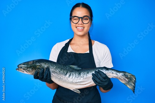 Young beautiful asian girl fishmonger selling fresh raw salmon winking looking at the camera with sexy expression, cheerful and happy face.