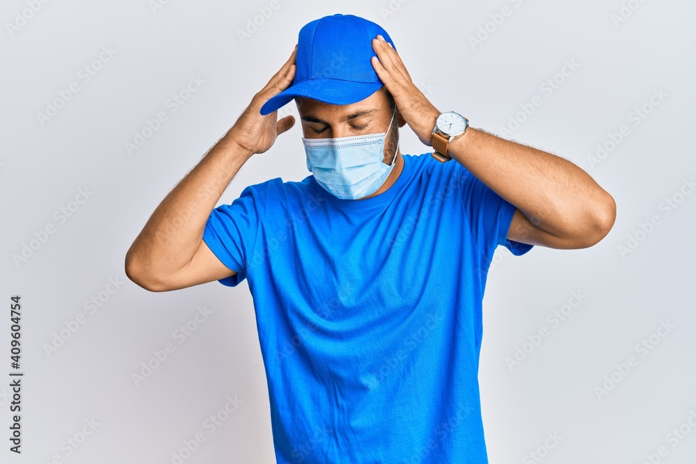 Handsome man with beard wearing courier uniform and medical mask with hand on head, headache because stress. suffering migraine.