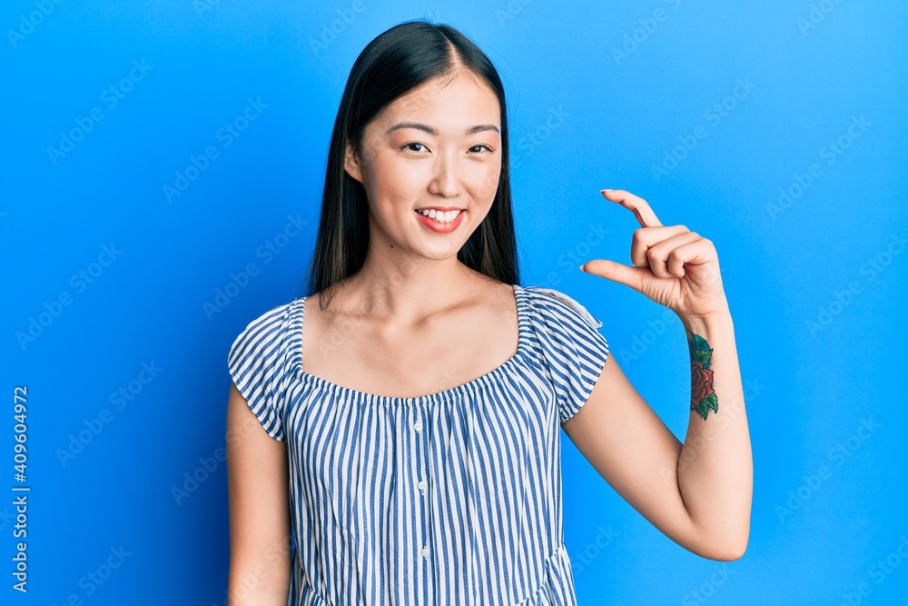 Young chinese woman wearing casual striped t-shirt smiling and confident gesturing with hand doing small size sign with fingers looking and the camera. measure concept. Young chinese woman wearing casual striped t-shirt smiling and confident gesturing with hand doing small size sign with fingers looking and the camera. measure concept.