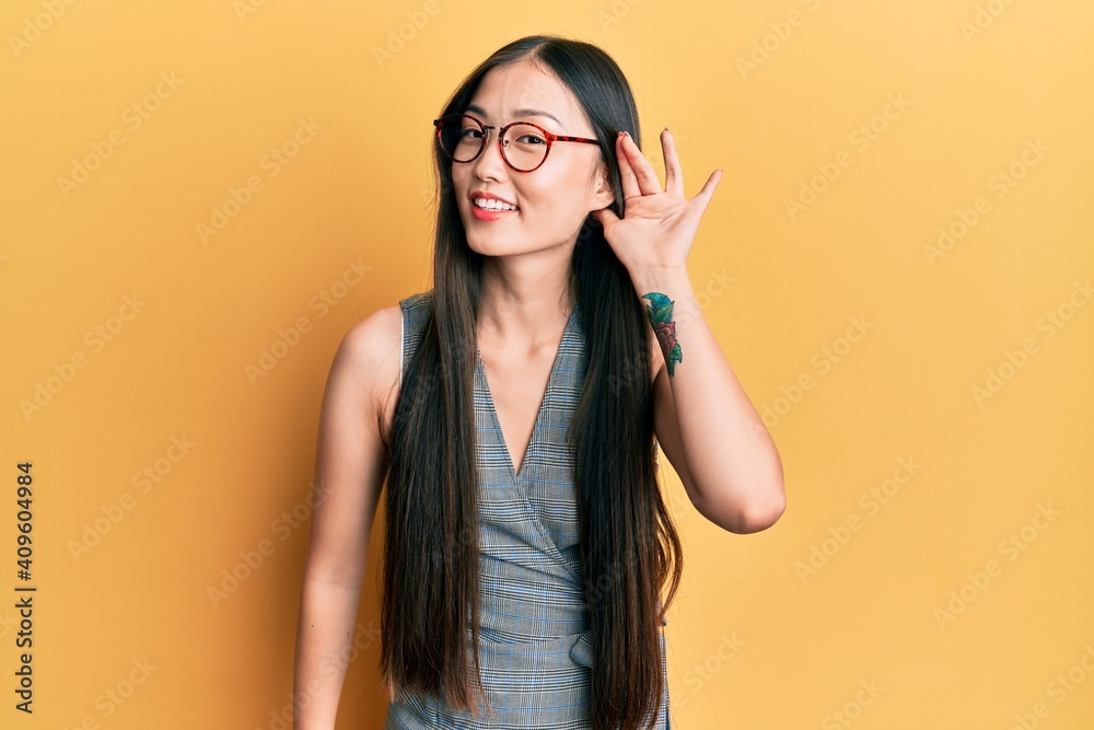 Young chinese woman wearing business dress and glasses smiling with hand over ear listening and hearing to rumor or gossip. deafness concept.