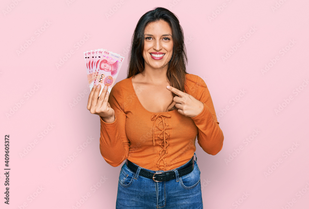 Beautiful hispanic woman holding 100 yuan chinese banknotes smiling happy pointing with hand and finger Beautiful hispanic woman holding 100 yuan chinese banknotes smiling happy pointing with hand and finger
