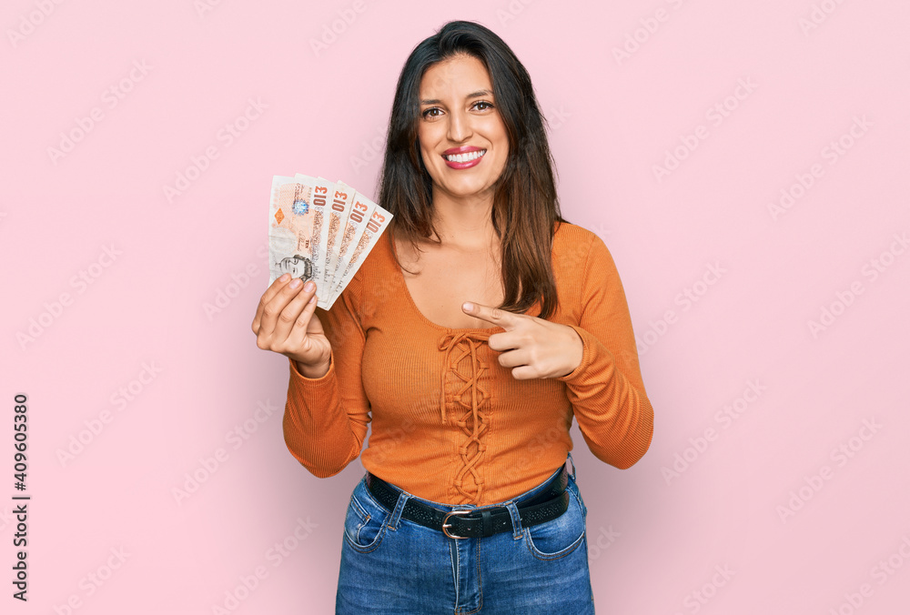 Beautiful hispanic woman holding 10 united kingdom pounds banknotes smiling happy pointing with hand and finger Beautiful hispanic woman holding 10 united kingdom pounds banknotes smiling happy pointing with hand and finger