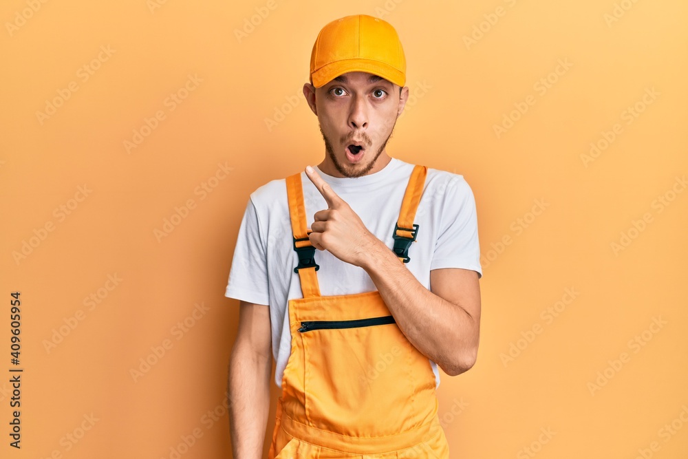 Hispanic young man wearing handyman uniform surprised pointing with finger to the side, open mouth amazed expression.