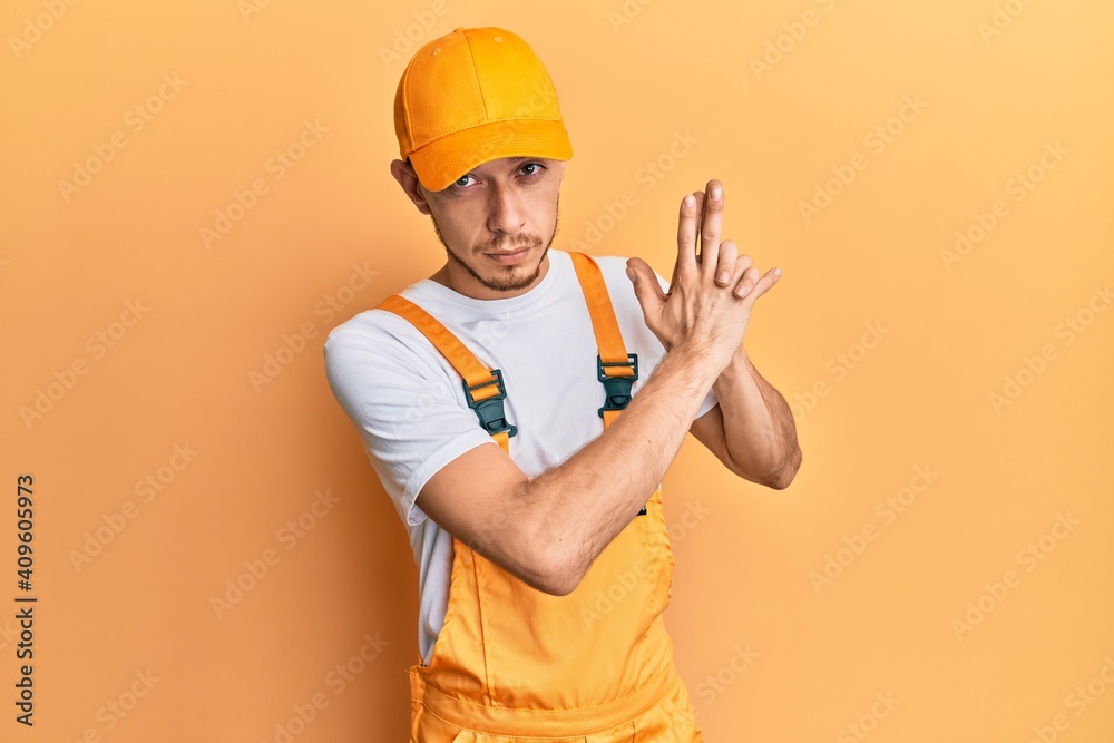 Hispanic young man wearing handyman uniform holding symbolic gun with hand gesture, playing killing shooting weapons, angry face
