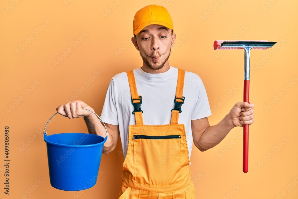 Hispanic young man wearing glass cleaner uniform and squeegee making fish face with mouth and squinting eyes, crazy and comical. Hispanic young man wearing glass cleaner uniform and squeegee making fish face with mouth and squinting eyes, crazy and comical.