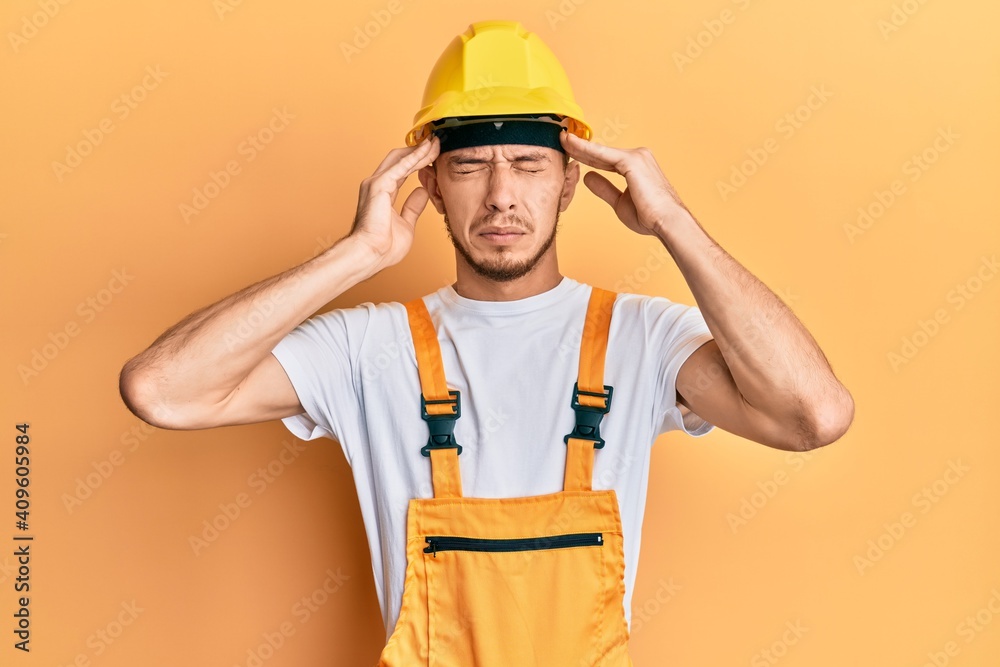 Hispanic young man wearing builder uniform and safety hardhat with hand on head, headache because stress. suffering migraine. Hispanic young man wearing builder uniform and safety hardhat with hand on head, headache because stress. suffering migraine.