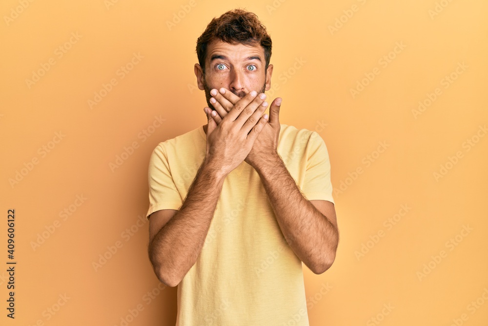 Handsome man with beard wearing casual yellow tshirt over yellow background shocked covering mouth with hands for mistake. secret concept. Handsome man with beard wearing casual yellow tshirt over yellow background shocked covering mouth with hands for mistake. secret concept.