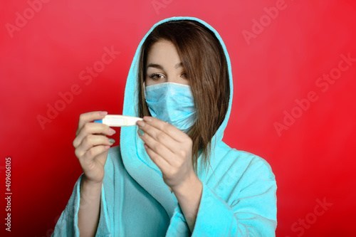 young woman in a mask on a red background holding a thermometer