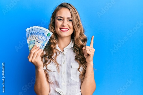 Young blonde girl holding south african 20 rand banknotes smiling with an idea or question pointing finger with happy face  number one