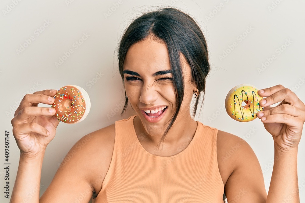 Young brunette woman holding tasty colorful doughnuts winking looking at the camera with sexy expression, cheerful and happy face. Young brunette woman holding tasty colorful doughnuts winking looking at the camera with sexy expression, cheerful and happy face.