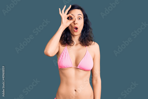 Young beautiful hispanic woman wearing bikini doing ok gesture shocked with surprised face, eye looking through fingers. unbelieving expression.