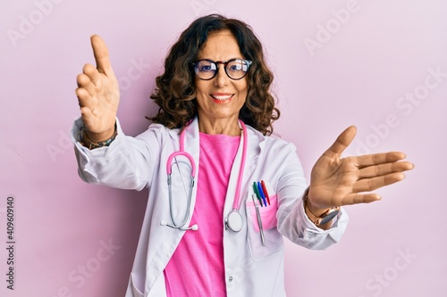 Middle age hispanic woman wearing doctor uniform and glasses looking at the camera smiling with open arms for hug. cheerful expression embracing happiness.