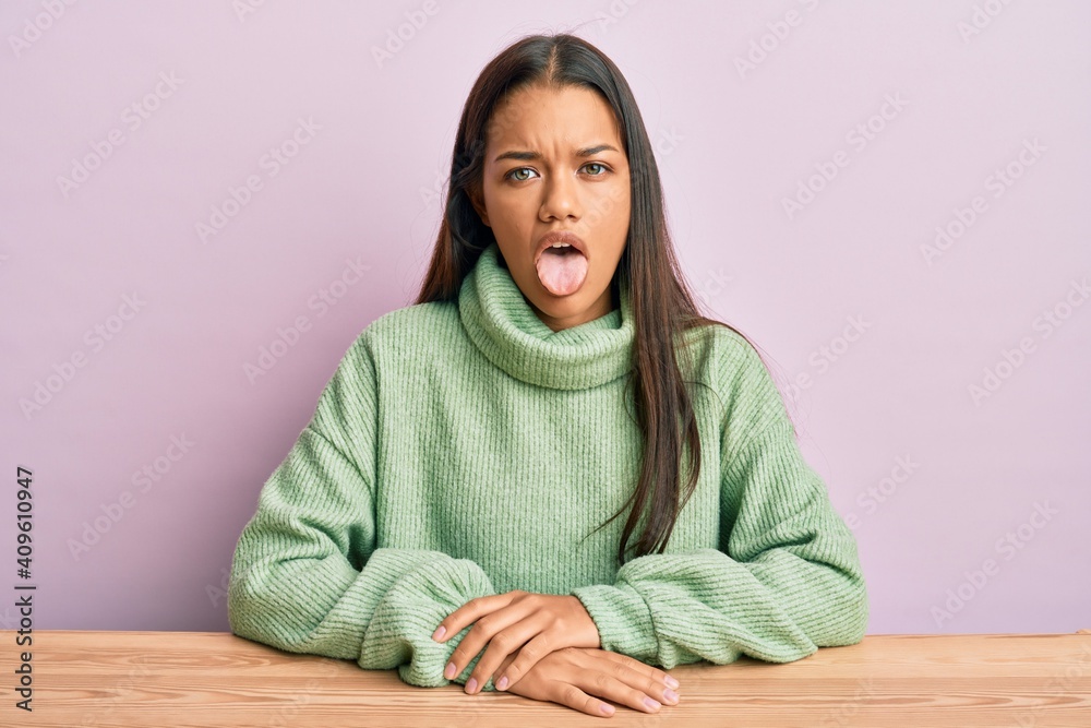 Beautiful hispanic woman wearing casual clothes sitting on the table sticking tongue out happy with funny expression. emotion concept. Beautiful hispanic woman wearing casual clothes sitting on the table sticking tongue out happy with funny expression. emotion concept.