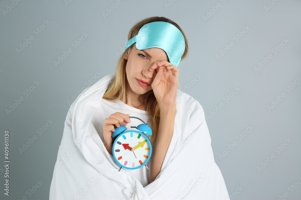 Young woman in sleeping mask wrapped with blanket holding alarm clock on grey background Young woman in sleeping mask wrapped with blanket holding alarm clock on grey background