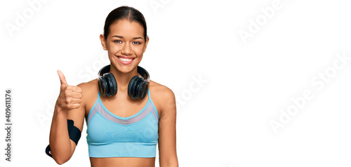 Beautiful hispanic woman wearing sportswear and headphones smiling happy and positive, thumb up doing excellent and approval sign