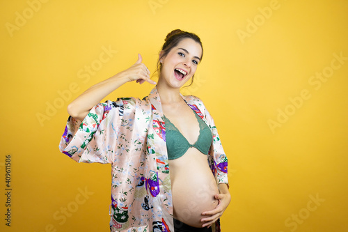 Young beautiful woman pregnant expecting baby wearing pajama over isolated yellow background smiling doing phone gesture with hand and fingers like talking on the telephone