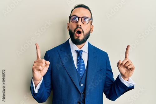 Young man with beard wearing business suit and tie amazed and surprised looking up and pointing with fingers and raised arms.