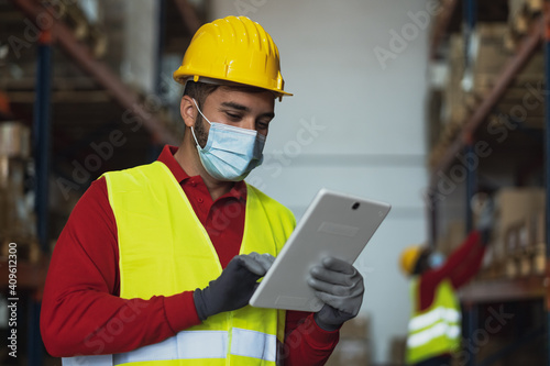 Young man working in warehouse doing inventory using digital tablet and loading delivery boxes plan while wearing face mask during corona virus pandemic - Logistic and industry concept