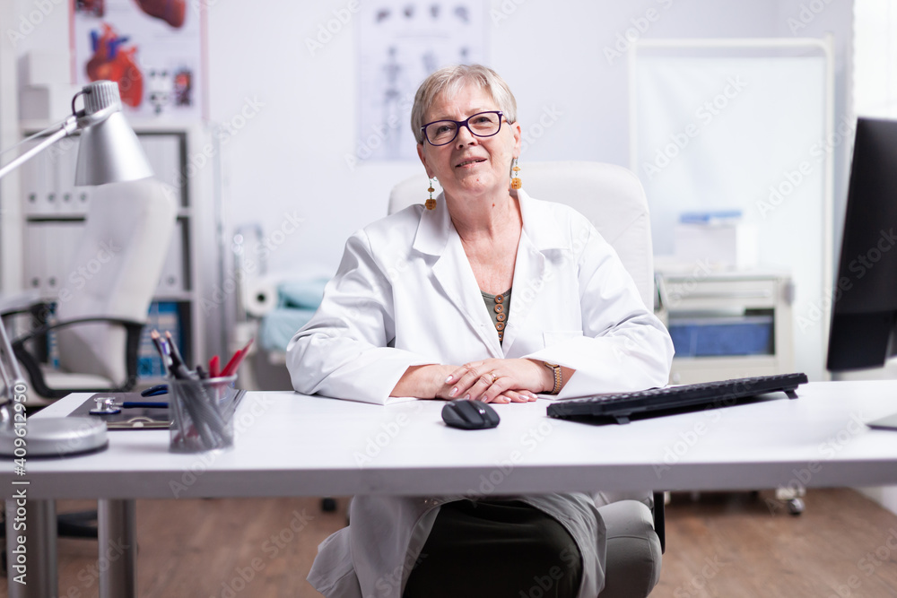 Senior medic smiling during video meeting with patients sitting at desk in medical clinic. POV of doctor speaking online in hospital room looking at camera wearing lab coat during telemedicine Senior medic smiling during video meeting with patients sitting at desk in medical clinic. POV of doctor speaking online in hospital room looking at camera wearing lab coat during telemedicine
