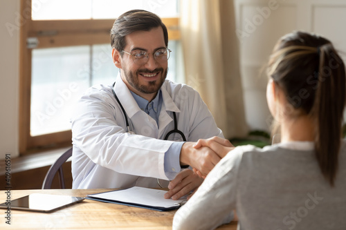 Smiling male doctor in white medical uniform shake hand greet with female patient at meeting in hospital. Happy man GP handshake woman client get acquainted or close deal at consultation in clinic.
