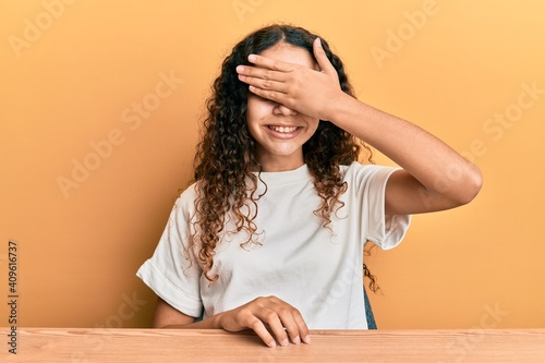 Teenager hispanic girl wearing casual clothes sitting on the table smiling and laughing with hand on face covering eyes for surprise. blind concept. © Krakenimages.com