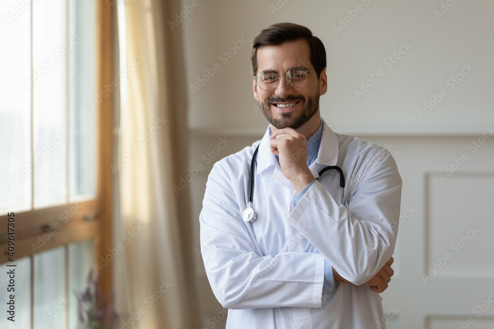 Portrait of smiling young Caucasian male doctor in white medical uniform look at camera show confidence success. Happy millennial man GP or pediatrician feel optimistic in private hospital or clinic.