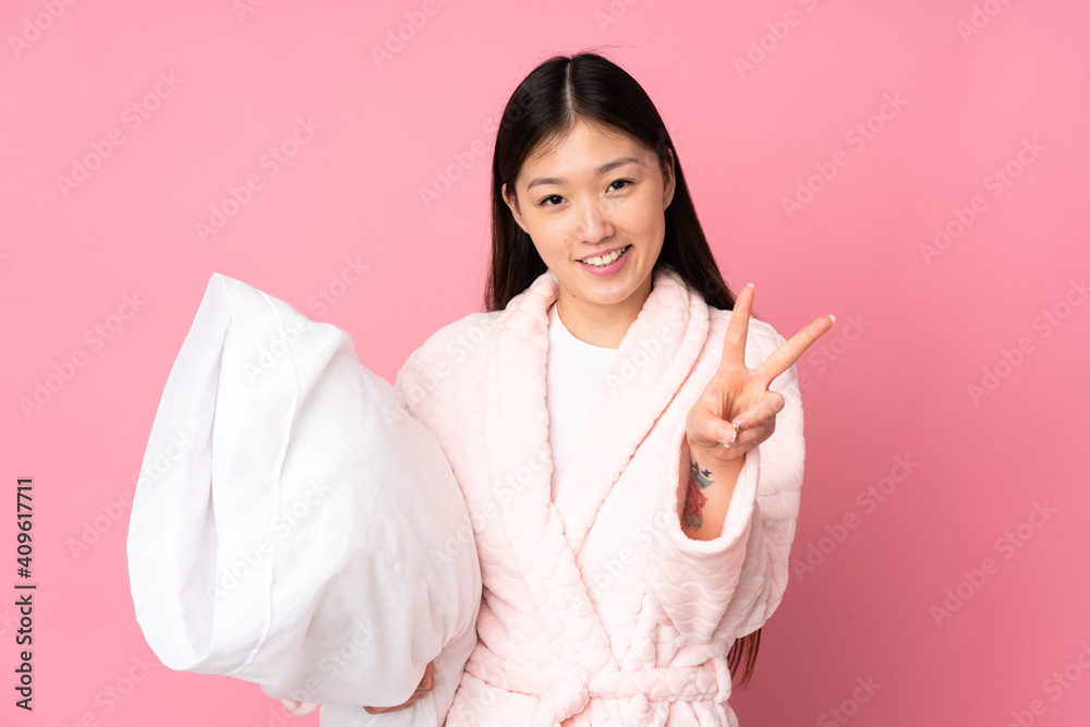 Young Asian woman in pajamas isolated on pink background smiling and showing victory sign Young Asian woman in pajamas isolated on pink background smiling and showing victory sign