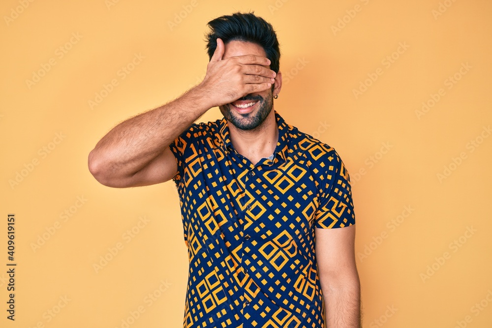 Handsome hispanic man with beard wearing 80s disco style shirt smiling and laughing with hand on face covering eyes for surprise. blind concept. Handsome hispanic man with beard wearing 80s disco style shirt smiling and laughing with hand on face covering eyes for surprise. blind concept.